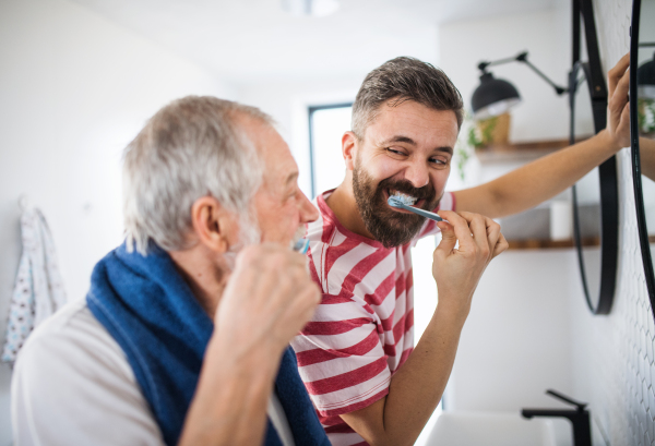 An adult hipster son and senior father brushing teeth in the bathroom indoors at home.