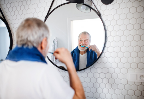 A senior man brushing teeth in bathroom indoors at home, looking in mirror. Copy space.