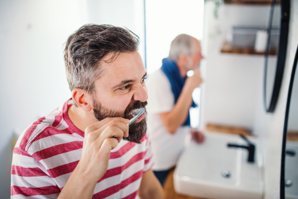 An adult hipster son and senior father brushing teeth in the bathroom indoors at home.