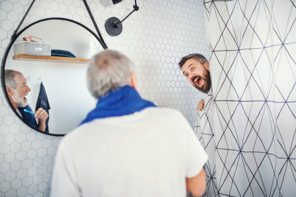 A cheerful adult hipster son and senior father in bathroom indoors at home, having fun.