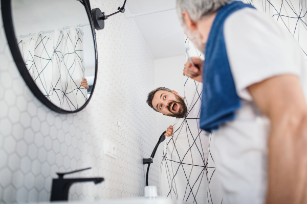 A cheerful adult hipster son and senior father in bathroom indoors at home, having fun.