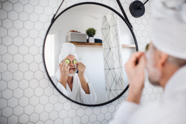 A senior man with cucumber on eyes indoors at home, looking in mirror. Copy space.