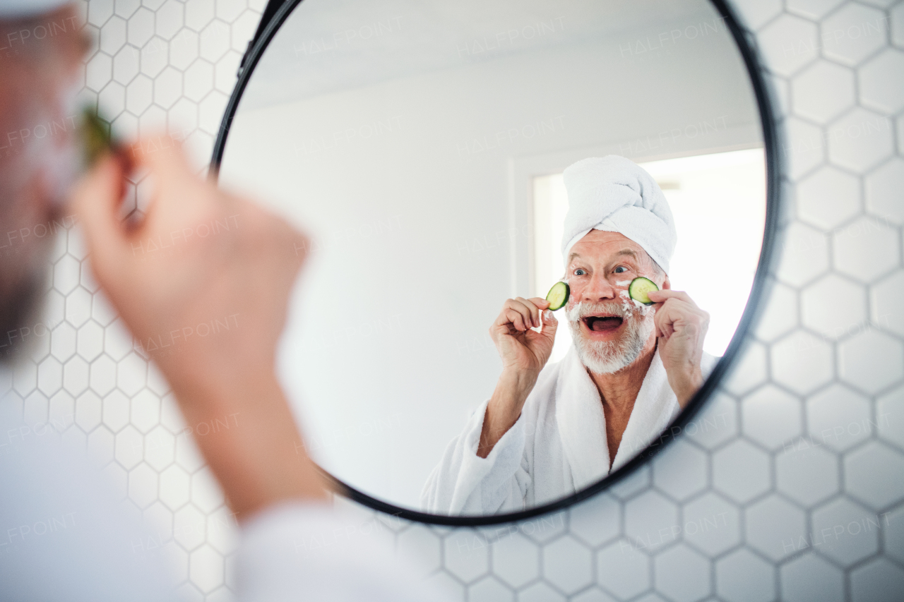 A senior man with cucumber on eyes indoors at home, looking in mirror. Copy space.