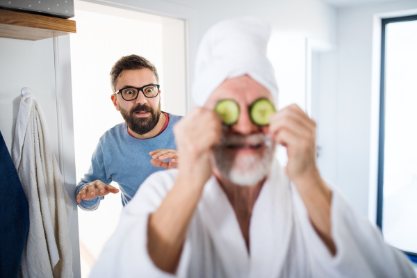 A cheerful adult hipster son and senior father in bathroom indoors at home, having fun.