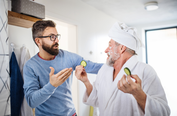 A cheerful adult hipster son and senior father in bathroom indoors at home, having fun.