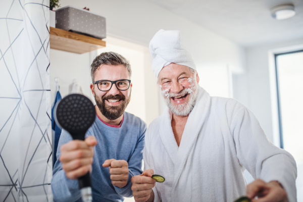 A cheerful adult hipster son and senior father in bathroom indoors at home, having fun.