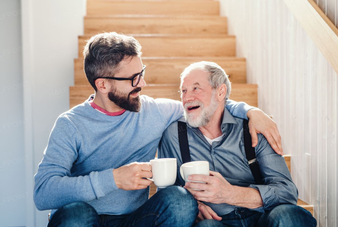 An adult hipster son and senior father with coffee sitting at the bottom of stairs indoors at home, talking.