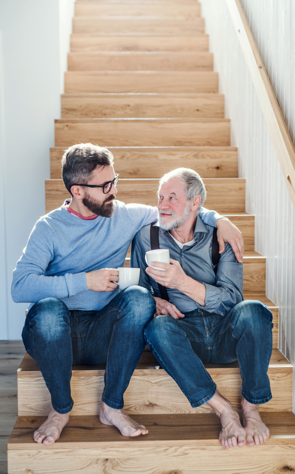 An adult hipster son and senior father with coffee sitting at the bottom of stairs indoors at home, talking.