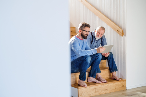 An adult hipster son and senior father with tablet sitting on stairs indoors at home.