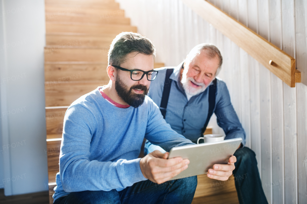 An adult hipster son and senior father with tablet sitting on stairs indoors at home.