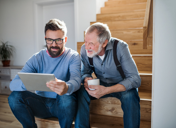 An adult hipster son and senior father with tablet sitting on stairs indoors at home.