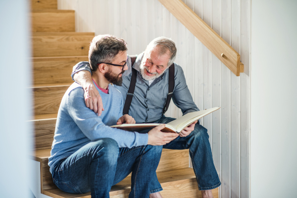 An adult hipster son and senior father sitting on stairs indoors at home, looking at photographs.