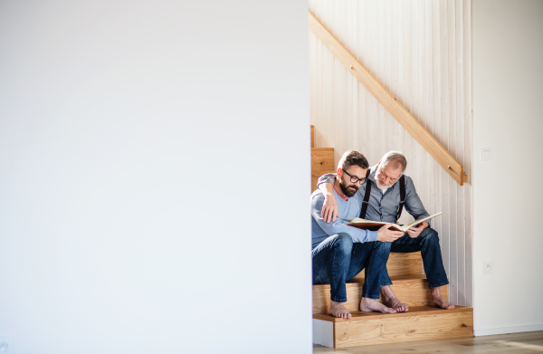 An adult hipster son and senior father sitting on stairs indoors at home, looking at photographs. Copy space.