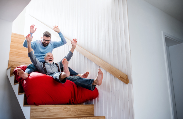 An adult hipster son and senior father sliding on stairs indoors at home, having fun.