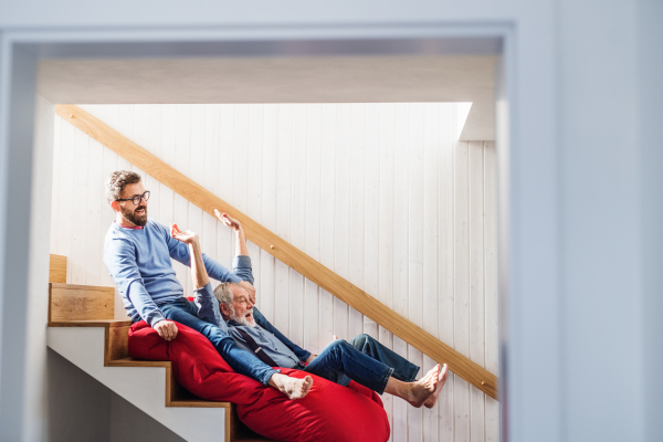 An adult hipster son and senior father sliding on stairs indoors at home, having fun.