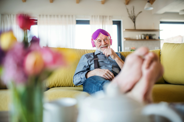 A senior man with headphones and eye mask sitting on sofa indoors at home, listening to music and relaxing.