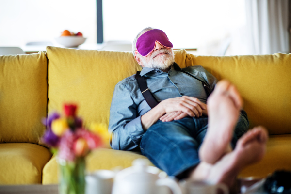 A senior man with eye mask lying on sofa indoors at home, relaxing.