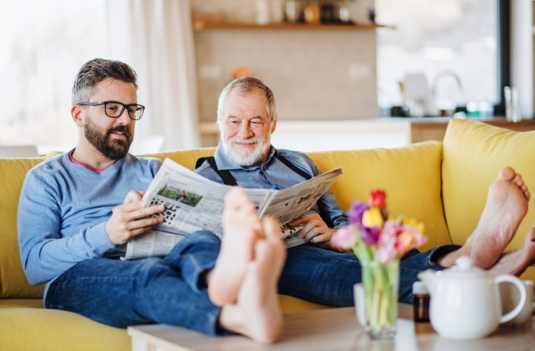 An adult hipster son and senior father with newspapers sitting on sofa indoors at home, talking.