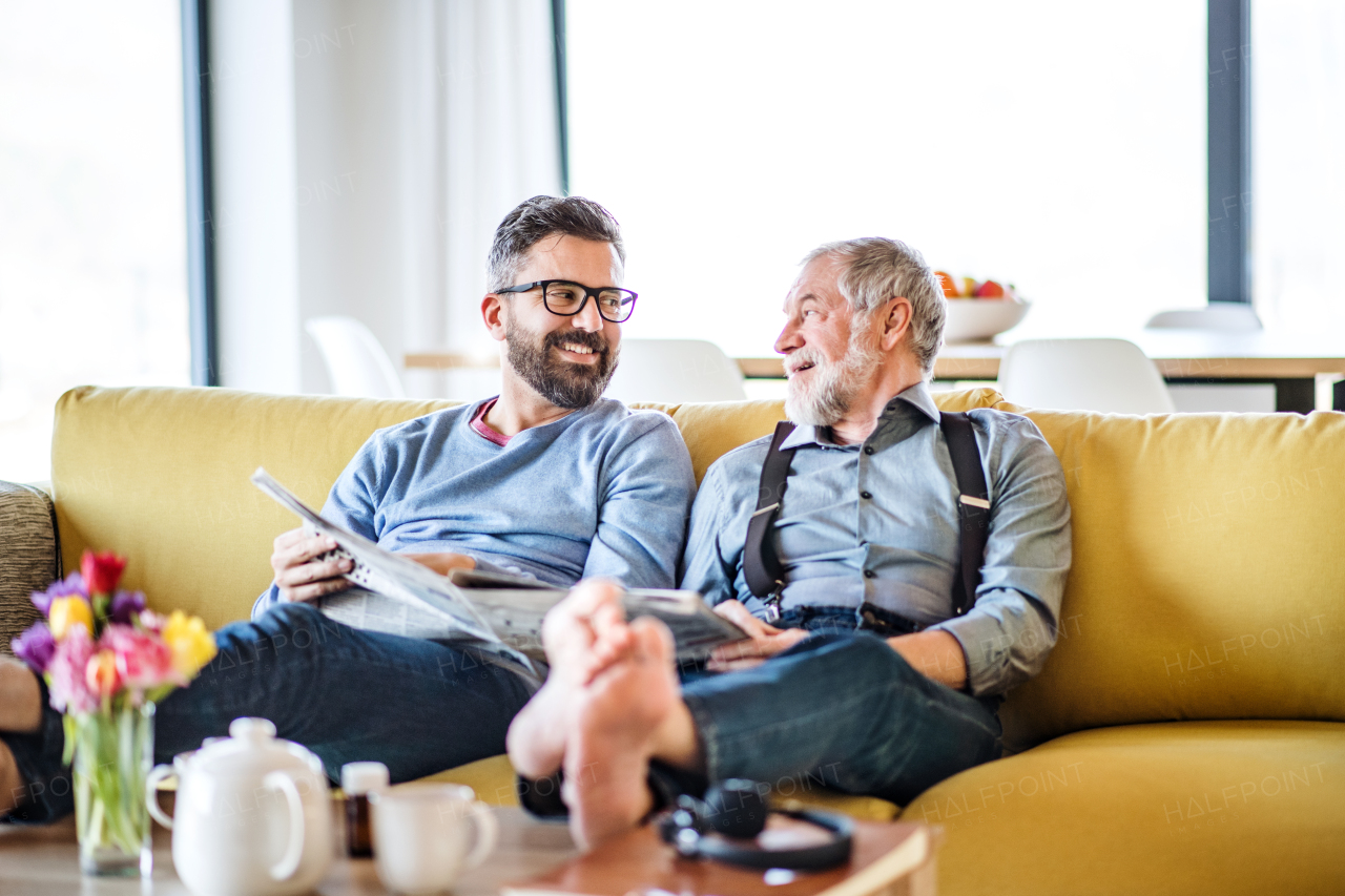 An adult hipster son and senior father with newspapers sitting on sofa indoors at home, talking.