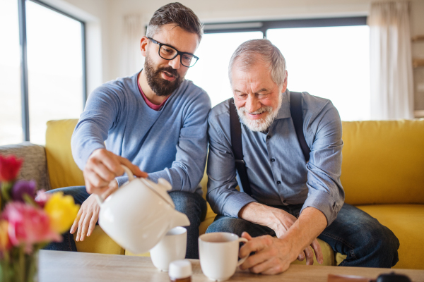 An adult hipster son and senior father sitting on sofa indoors at home, drinking tea.