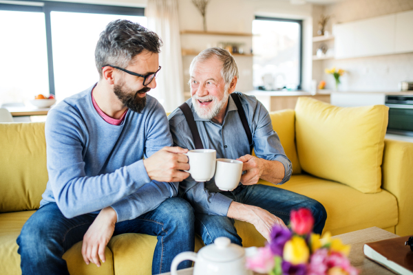 An adult hipster son and senior father sitting on sofa indoors at home, drinking tea.