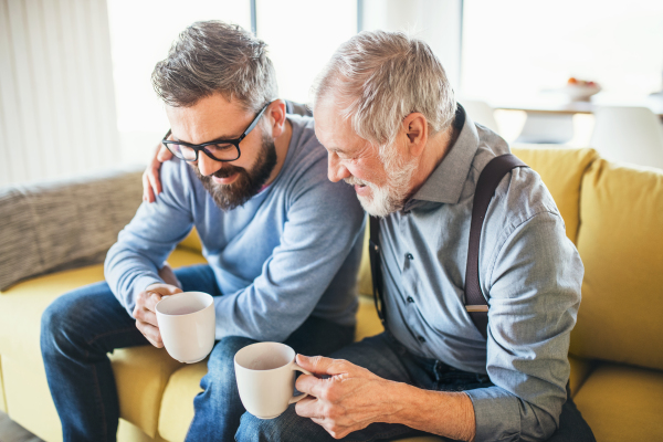 An adult hipster son and senior father with coffee sitting on sofa indoors at home, talking.