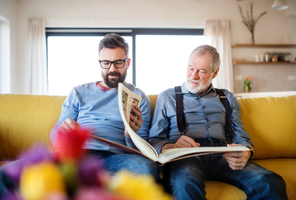 An adult hipster son and senior father with photo album sitting on sofa indoors at home, talking.