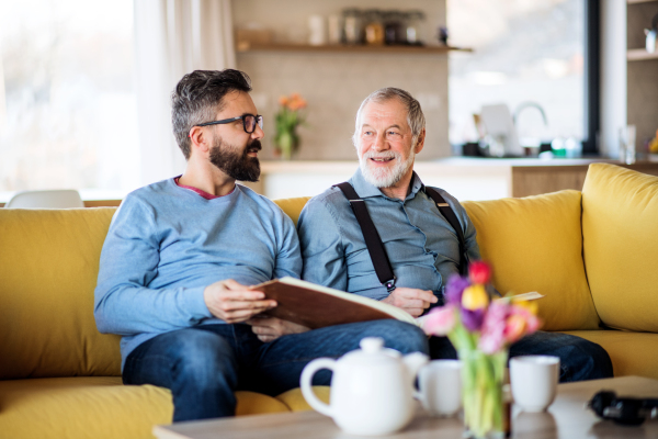 An adult hipster son and senior father with photo album sitting on sofa indoors at home, talking.