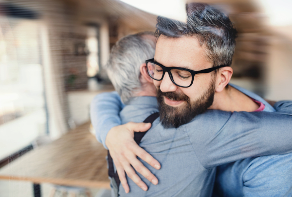 An adult hipster son with glasses and senior father indoors at home, hugging.