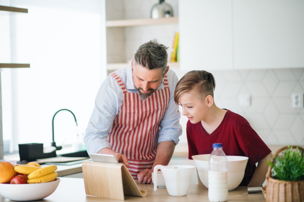 A mature father and small son with tablet indoors in kitchen, making pancakes.