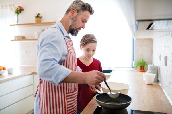 A mature father with small son indoors in kitchen, making pancakes.