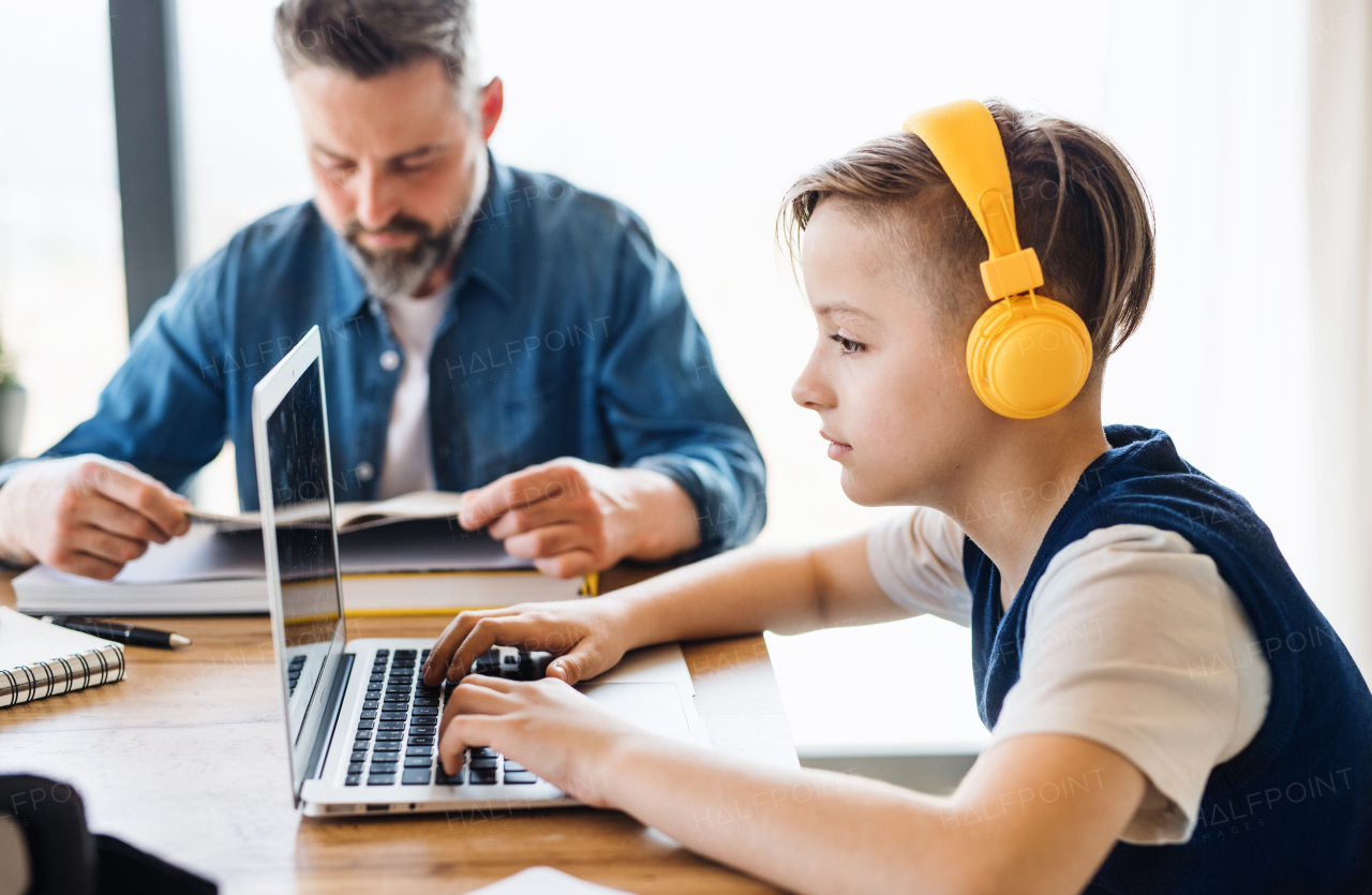 A mature father with small son sitting at table indoors, using laptop and headphones.