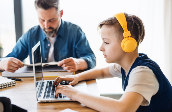 A mature father with small son sitting at table indoors, using laptop and headphones.