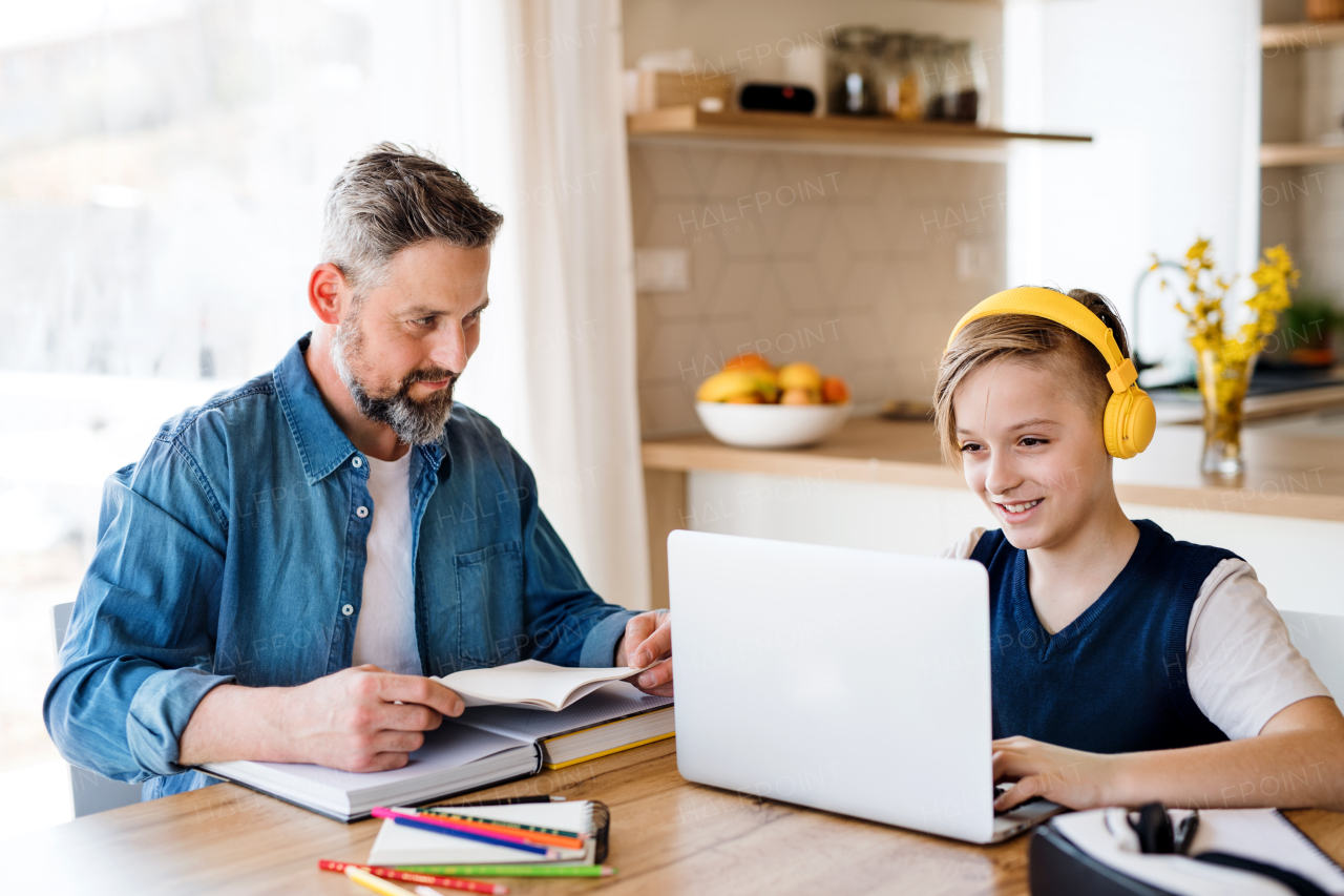 A mature father with small son sitting at table indoors, using laptop and headphones.