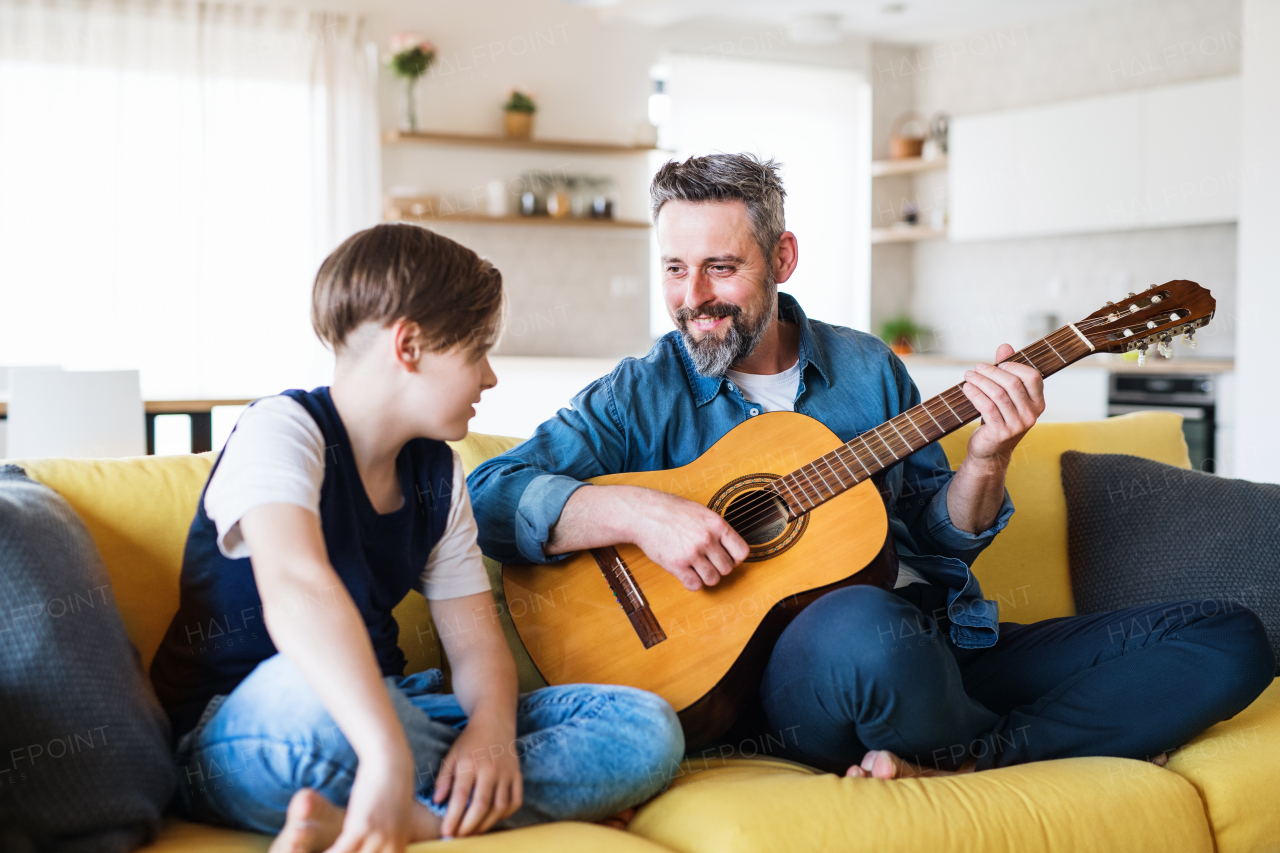 A mature father with small son sitting on sofa indoors, playing guitar.