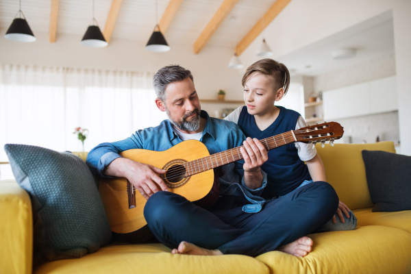 A mature father with small son sitting on sofa indoors, playing guitar..