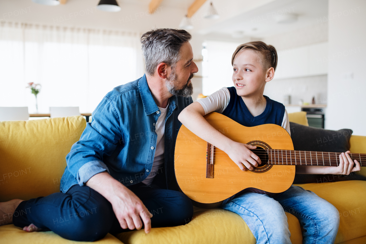 A mature father with small son sitting on sofa indoors, playing guitar.