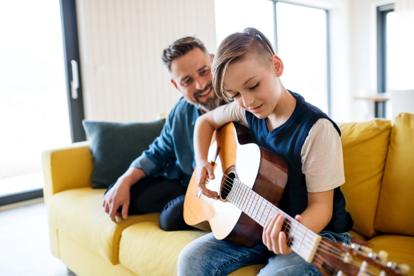 A mature father with small son sitting on sofa indoors, playing guitar.