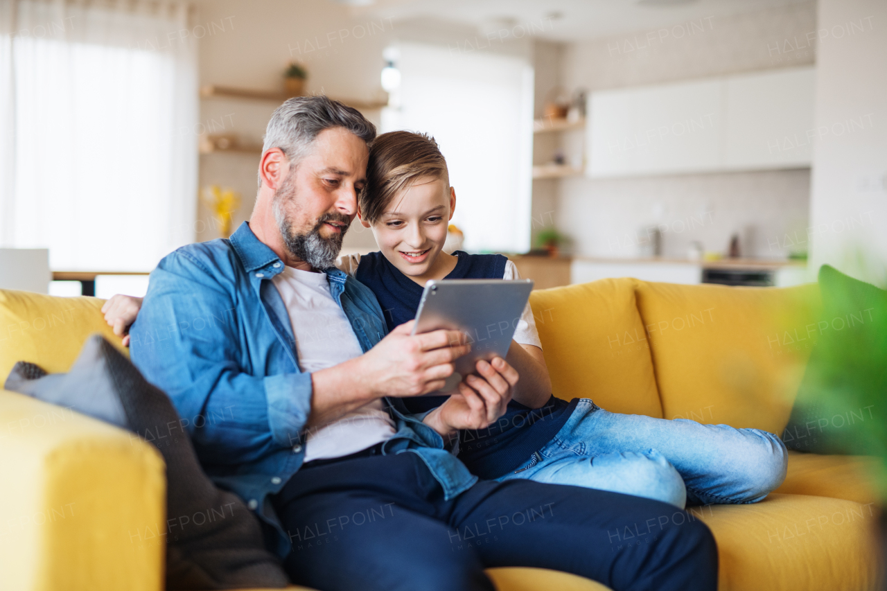 A mature father with small son sitting on sofa indoors, using tablet.