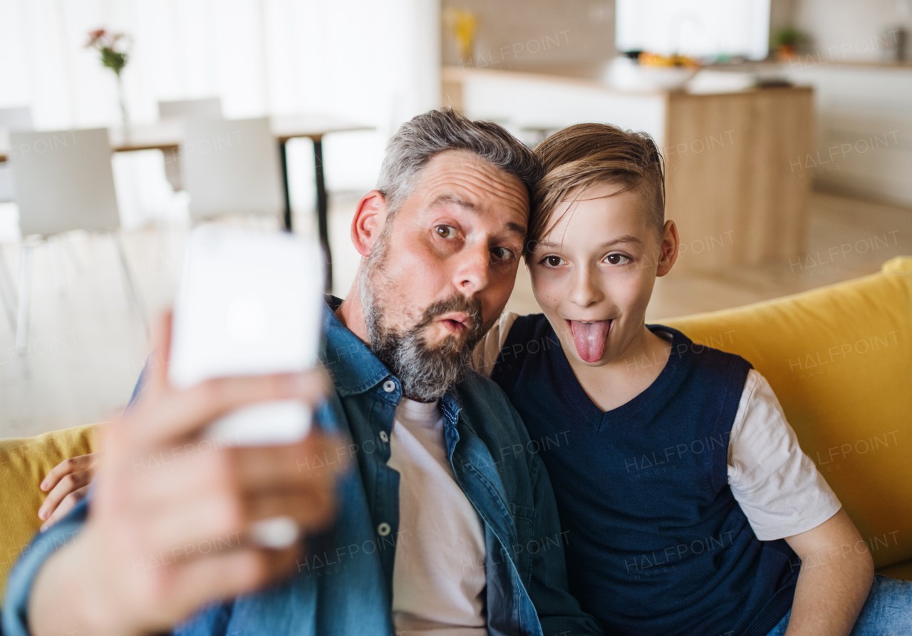 Mature father with small son sitting on sofa indoors, grimacing when taking selfie.