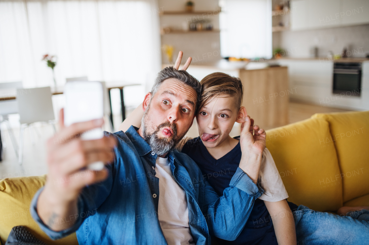 Mature father with small son sitting on sofa indoors, grimacing when taking selfie.