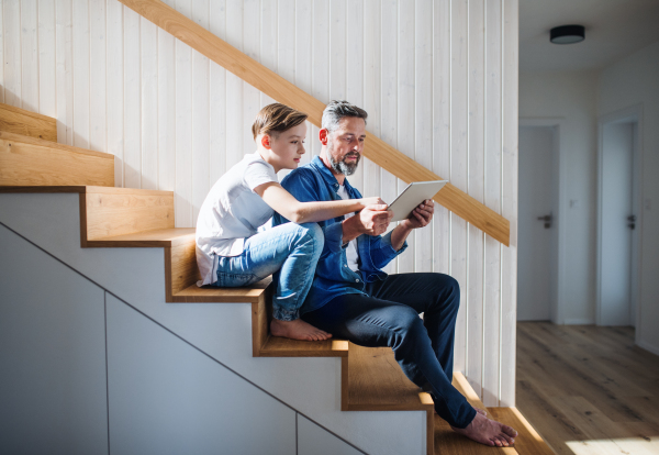 A mature father with small son sitting on the stairs indoors, using tablet.