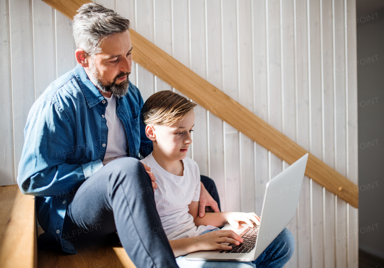 A mature father with small son sitting on the stairs indoors, using laptop.