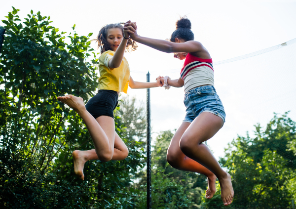 Low angle view of cheerful young teenager girls friends outdoors in garden, jumping on trampoline.