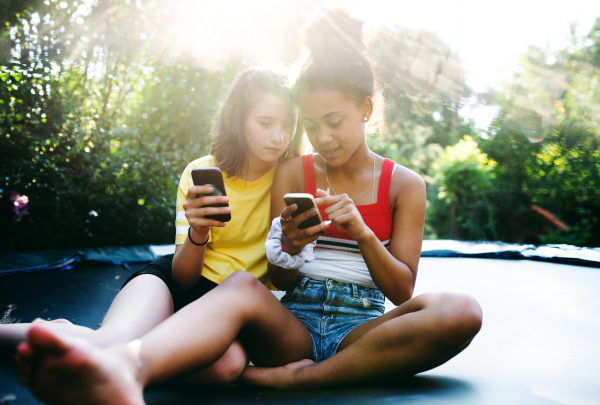 Front view of cheerful young teenager girls friends outdoors in garden, using smartphone.