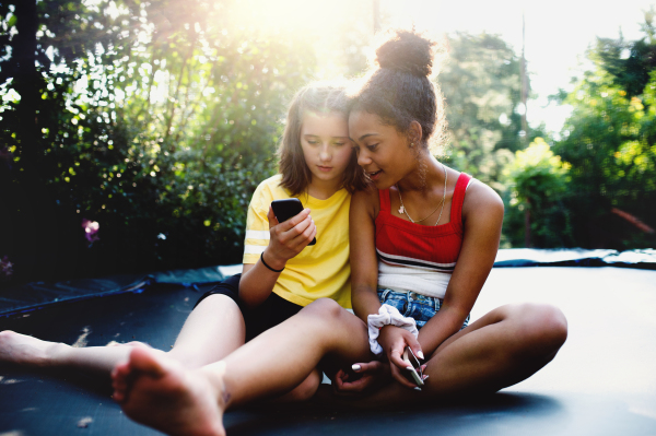 Front view of cheerful young teenager girls friends outdoors in garden, using smartphone.