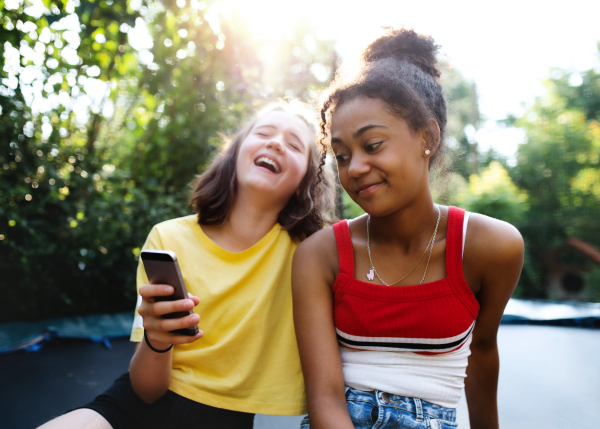 Front view of cheerful young teenager girls friends outdoors in garden, using smartphone.