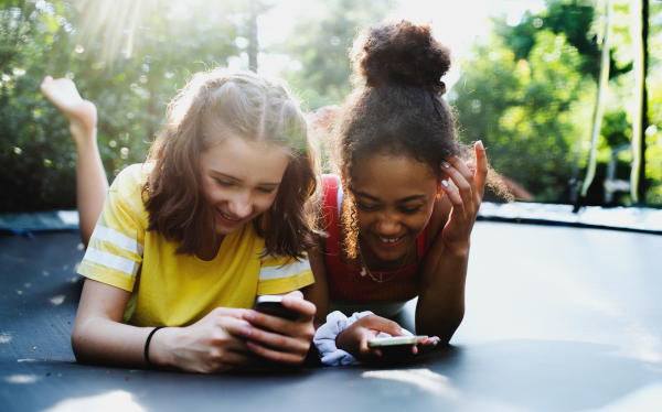 Front view of cheerful young teenager girls friends outdoors in garden, using smartphone.