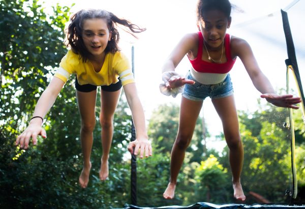 Front view of cheerful young teenager girls friends outdoors in garden, jumping on trampoline.