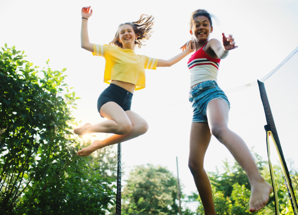 Low angle view of cheerful young teenager girls friends outdoors in garden, jumping on trampoline.